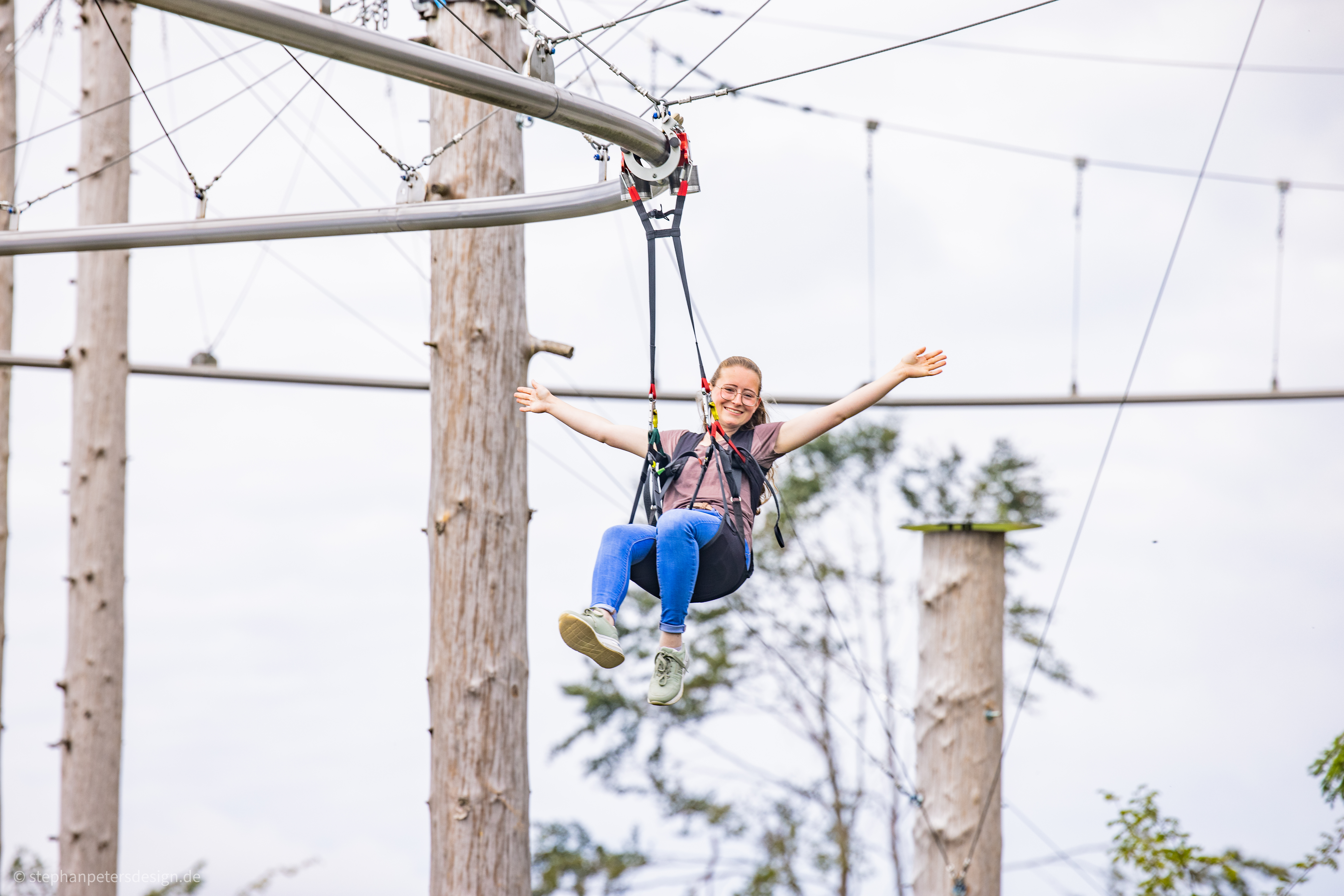 Eine Frau hängt in einem Sitzgurt an einer Metallschiene und breitet die Arme aus.