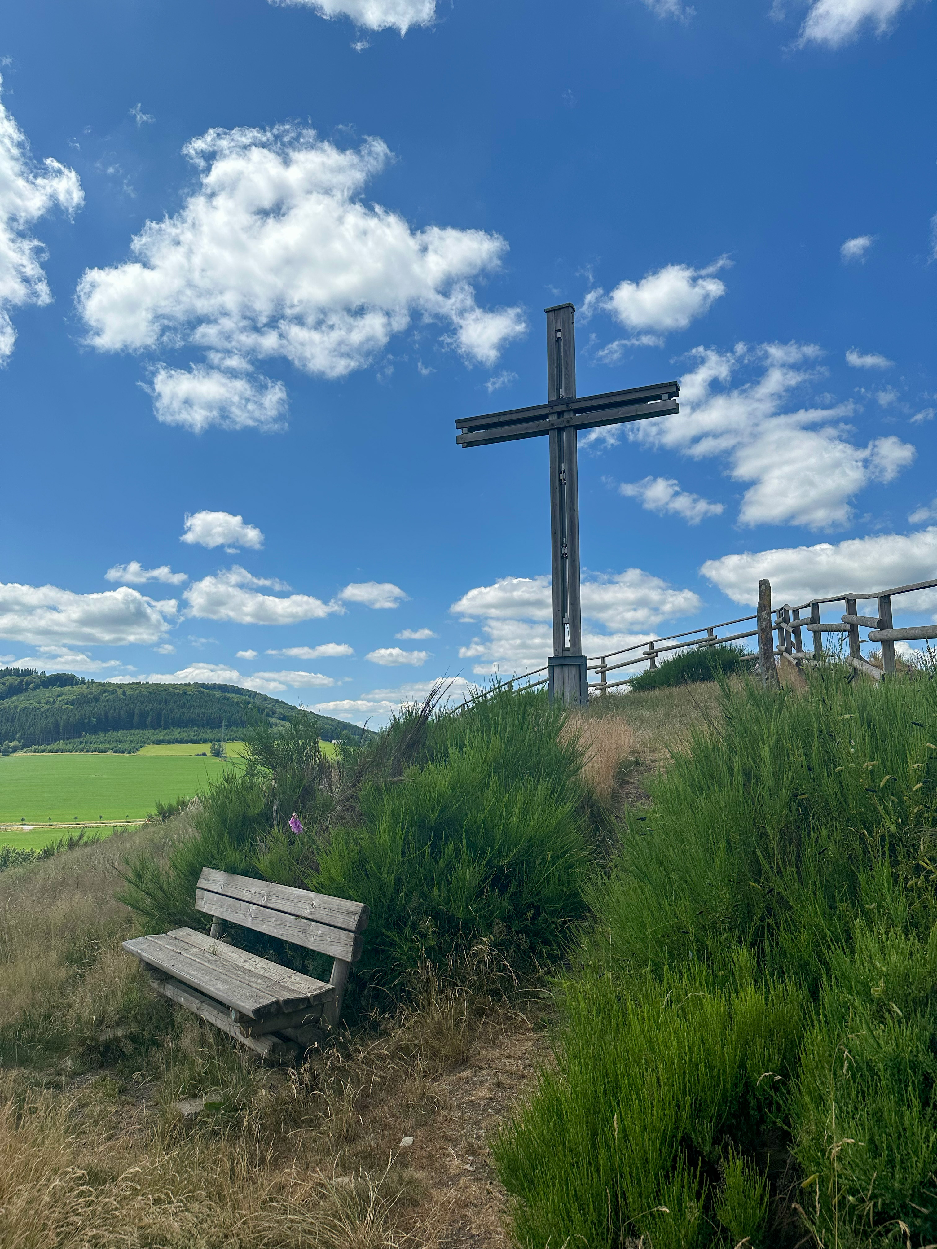 Ein großes Holzkreuz auf einem begrünten Hügel, davor eine Bank. Ein großes Holzkreuz auf einem begrünten Hügel, davor eine Bank.