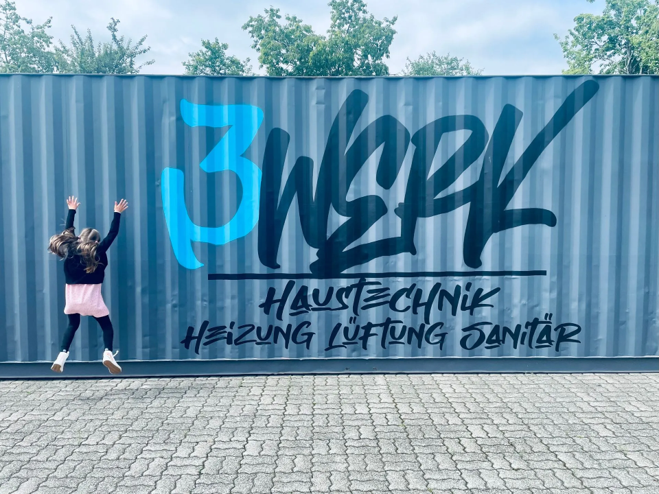 Springendes Mädchen vor grauem Hochseecontainer mit großer 3Werk Haustechnik Beschriftung.png
