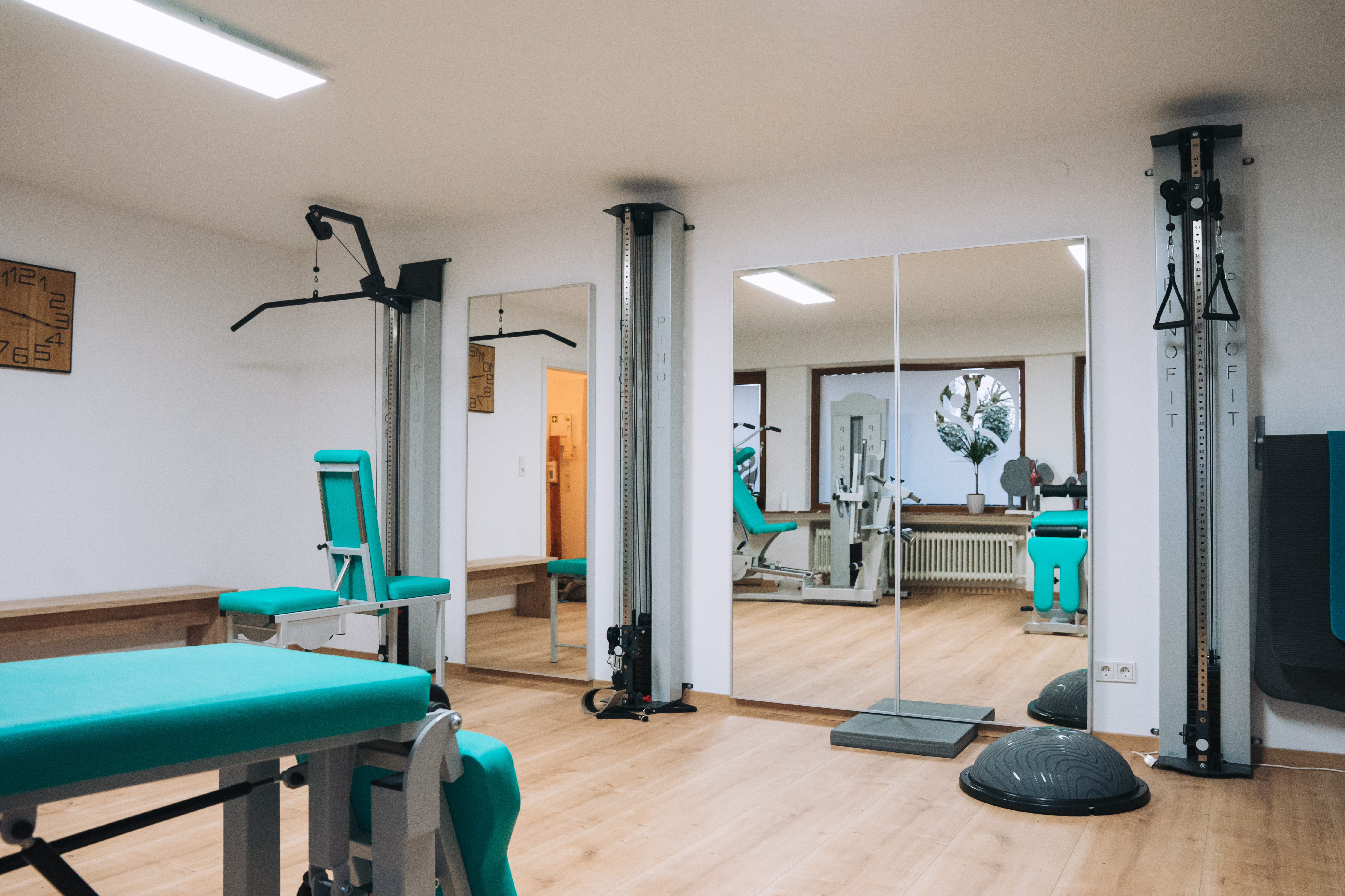 Heller Trainingsraum mit Geräten für Kraft- und Bewegungsübungen. Im Raum stehen mehrere Fitnessgeräte mit türkisfarbenen Polstern, große Spiegel an der Wand und ein Holzboden. Durch die Fenster fällt Tageslicht, und die Atmosphäre wirkt ordentlich und ruhig.jpg