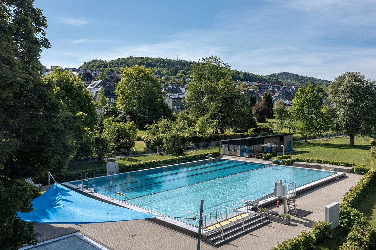 Ein Freibad mit großem Schwimmbecken umgeben von einer Liegewiese und Wald.